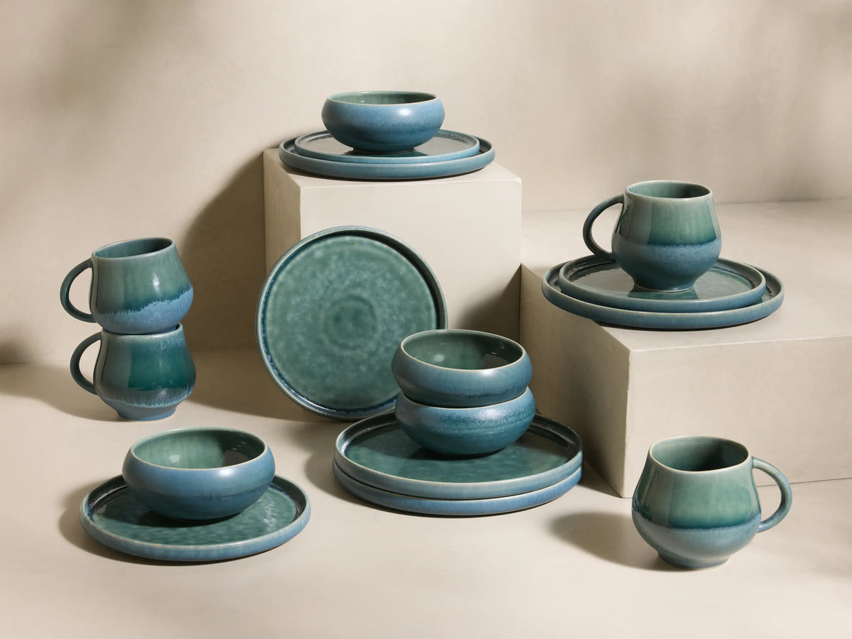 Full view of the blue-green reactive glaze Rhoswen Dinnerware with Cereal Bowls (Set of 16), displayed on neutral blocks.