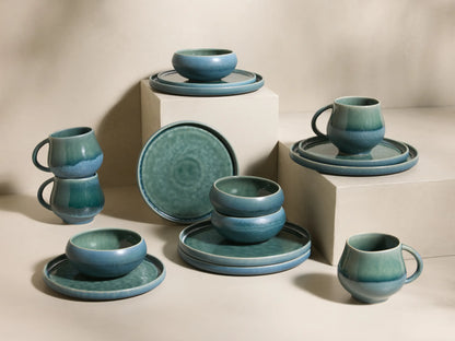 Full view of the blue-green reactive glaze Rhoswen Dinnerware with Cereal Bowls (Set of 16), displayed on neutral blocks.