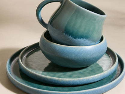 Full view of the blue-green reactive glaze Rhoswen Dinnerware with Cereal Bowls (Set of 16), displayed on neutral blocks.