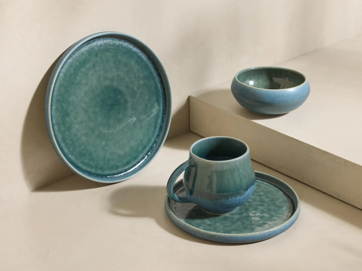 Close-up of stacked Rhoswen Dinnerware With Cereal Bowls, featuring a blue-green reactive glaze mug, cereal bowl, and plates.