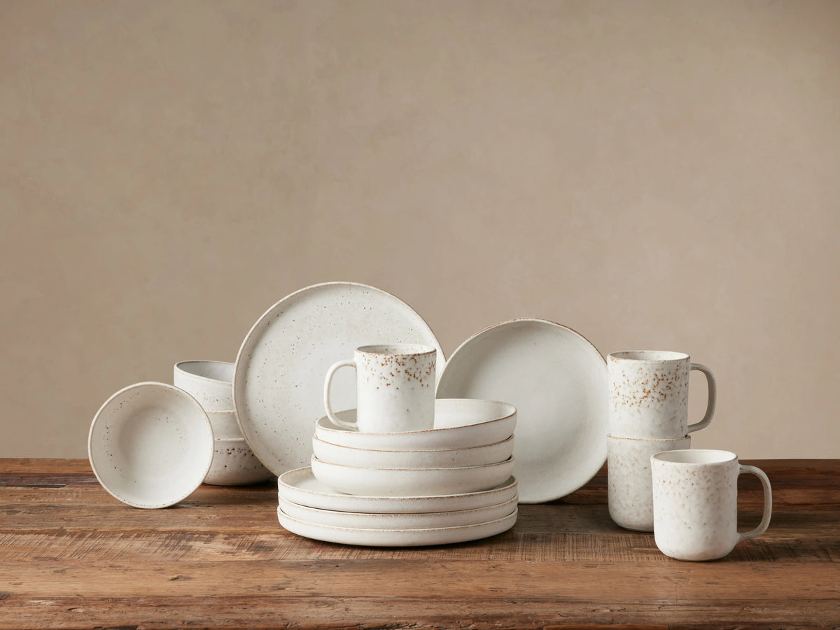 Ronan 16-pc white stoneware dinnerware set, including plates, bowls, and mugs, with speckled reactive glaze on a wooden table.