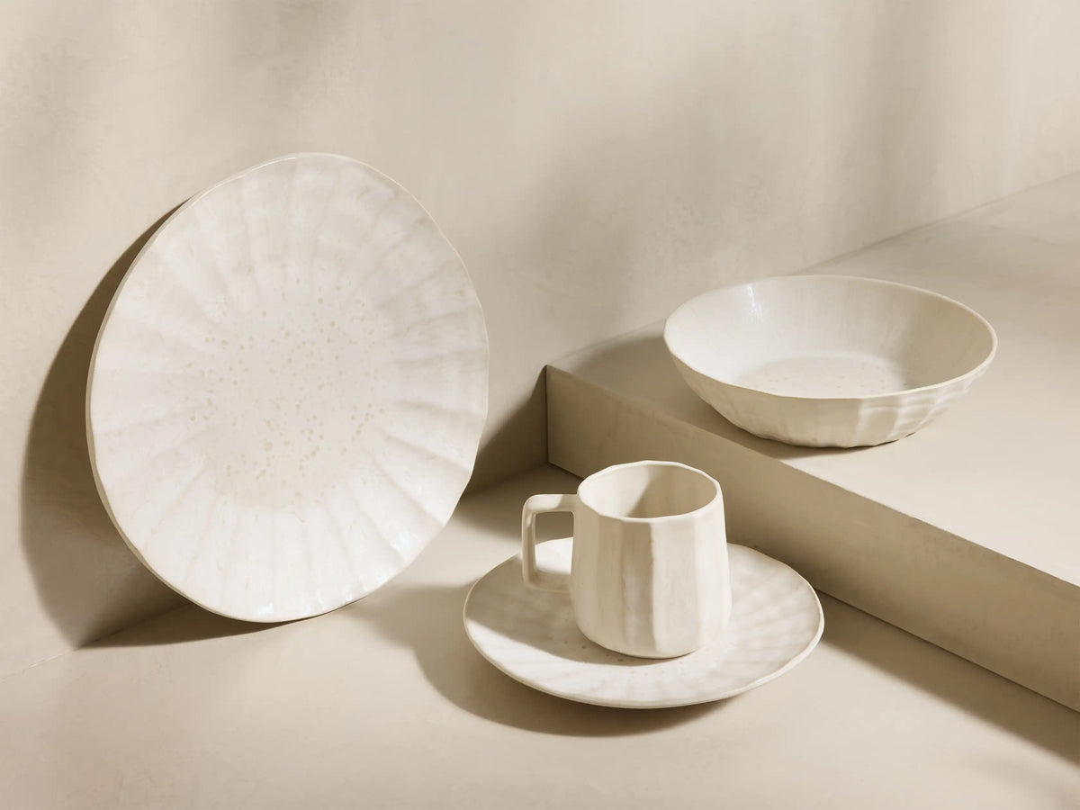 Close-up of creamy white Saskia Dinnerware, featuring a faceted mug, a speckled pasta bowl, and two stacked marbled plates.