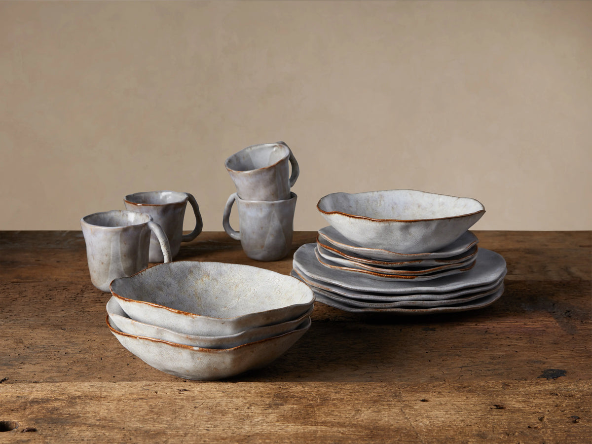 Handcrafted Skye Dinnerware set: stacked gray/white pasta bowls, plates, and mugs with rustic brown trim on a wooden table.