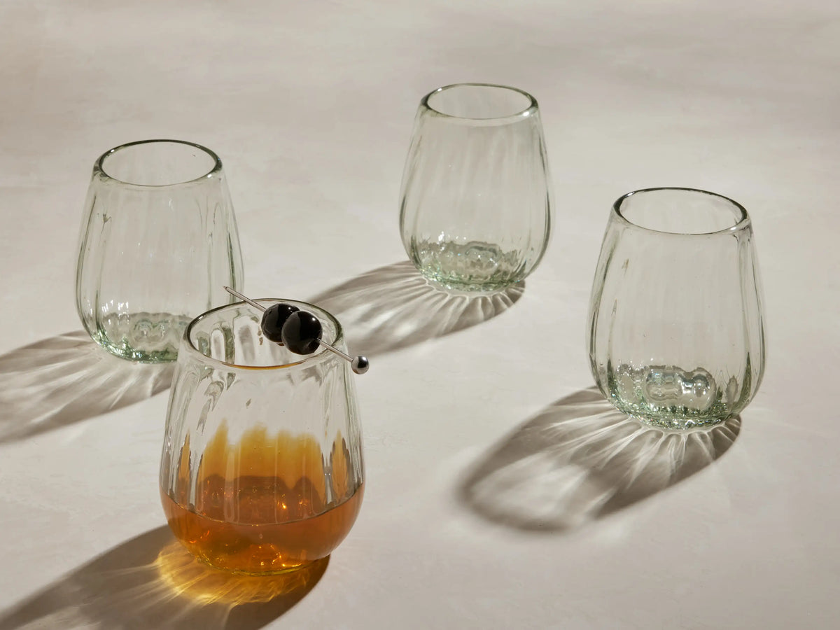 Four clear, textured Seema Double Old-Fashioned Glasses on a light surface. One holds amber liquid with two dark cherries.