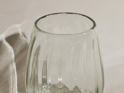Four clear, textured Seema Double Old-Fashioned Glasses on a light surface. One holds amber liquid with two dark cherries.