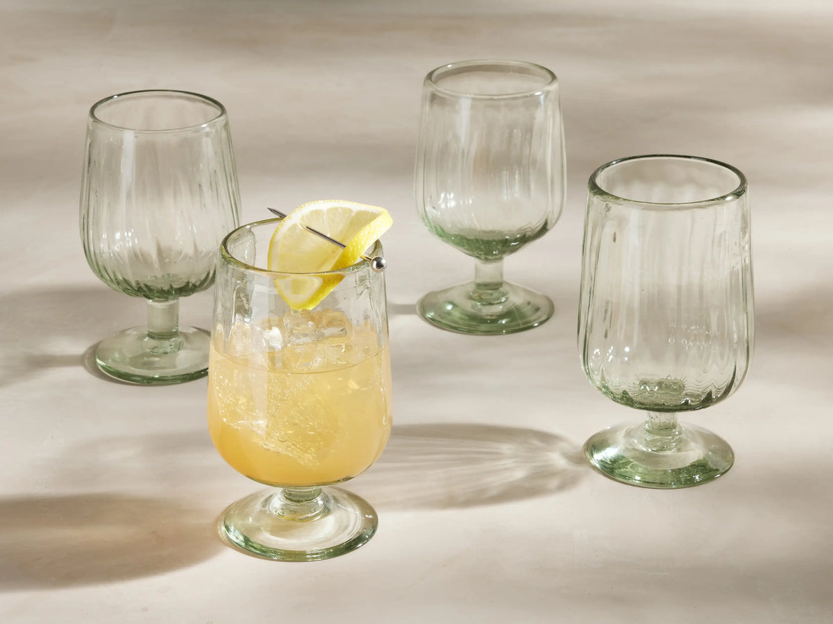 Four ribbed Seema Clear Goblet Glasses with a green tint. One glass in front holds a yellow drink with ice & lemon slice.