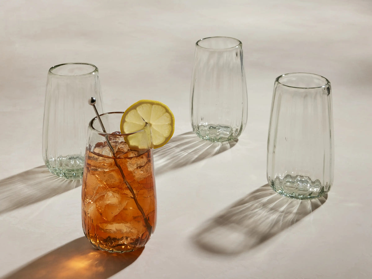 Seema clear highball glasses, set of 4. One glass contains iced amber drink with lemon & stirrer. Textured, recycled glass.