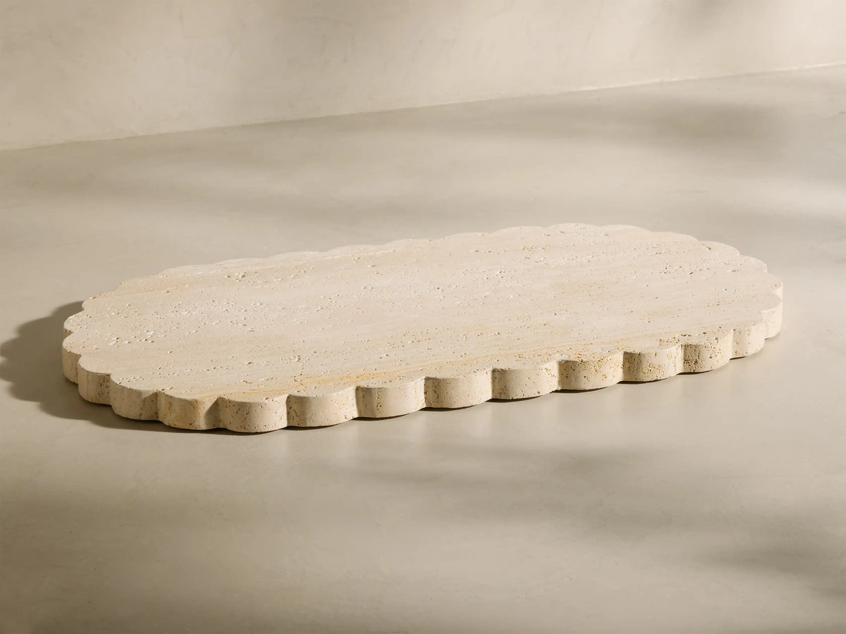 Angled view of the beige and ivory-toned travertine Sylvaine Oval Board, featuring scalloped edges and visible stone texture.