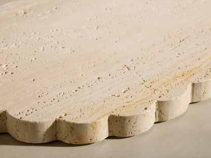 Angled view of the beige and ivory-toned travertine Sylvaine Oval Board, featuring scalloped edges and visible stone texture.