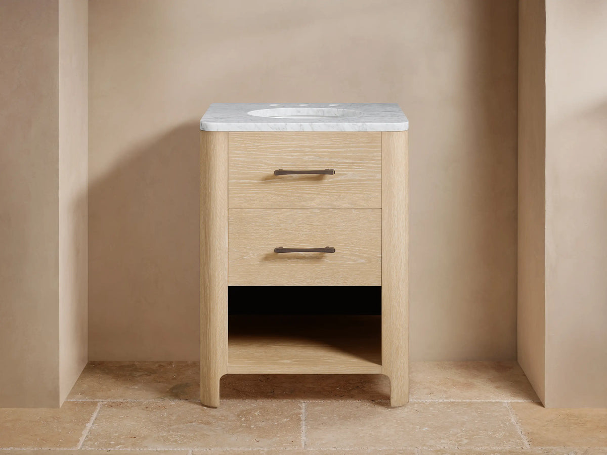 Calista Powder Vanity: Light oak wood vanity with white marble top, oval sink, dark faucet, 2 drawers with bronze pulls, open shelf.