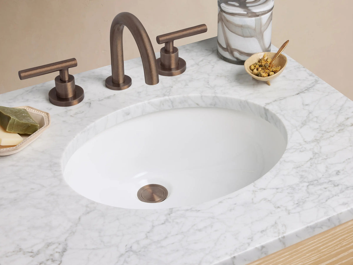 Calista Powder Vanity, light oak wood with white marble top, oval sink, two drawers with dark pulls, and an open shelf.
