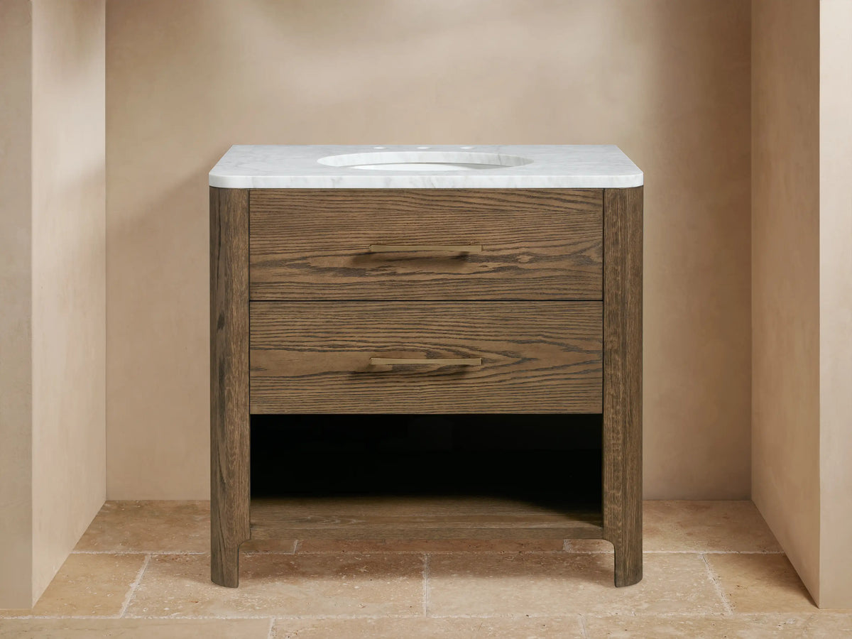 Calista Single Vanity in oak wood with two faux drawers, Carrara marble top, oval sink, brass hardware, and open shelf below.