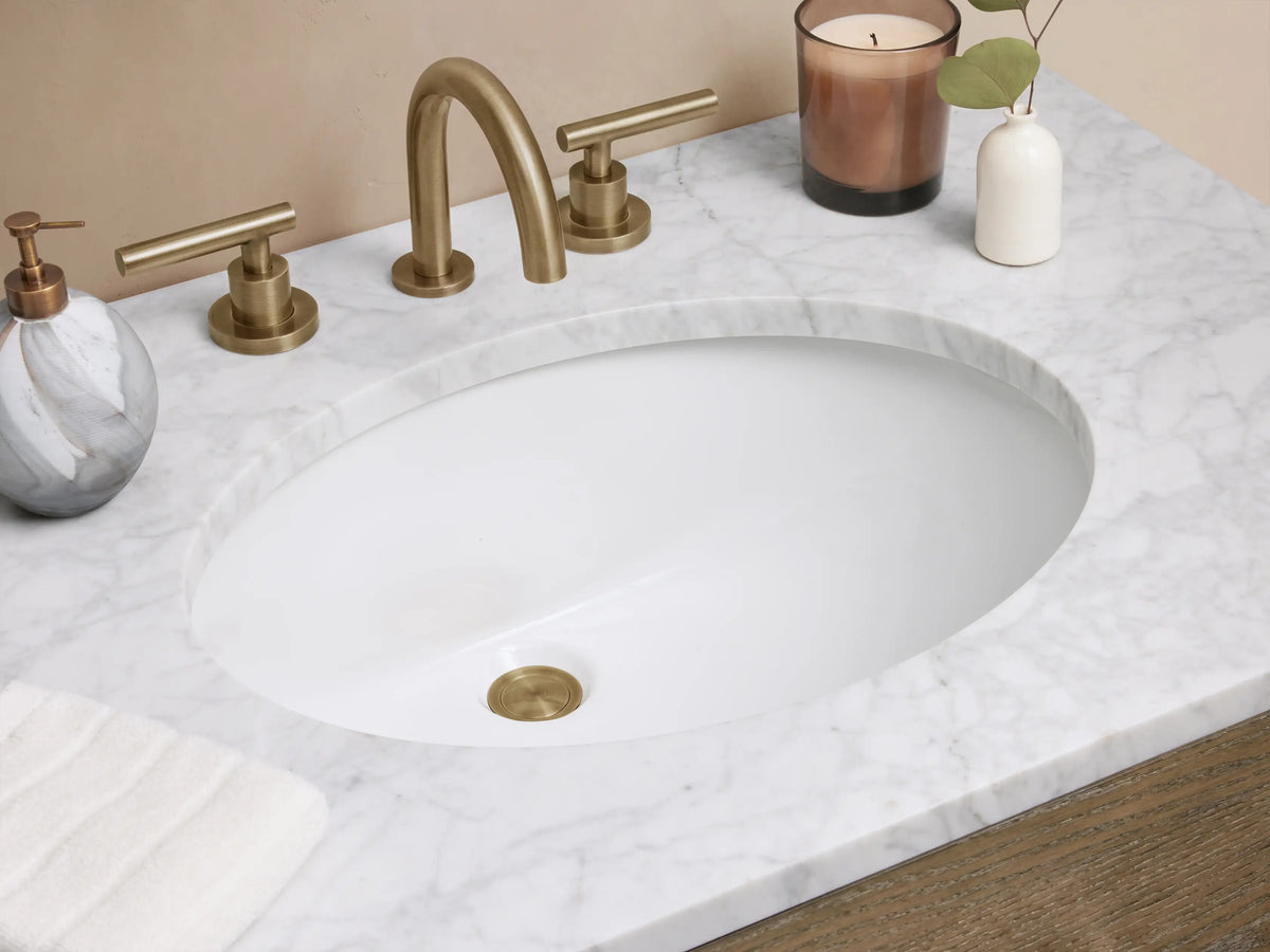 Calista Single Vanity with oak wood body, two drawers, stone top, oval sink, polished handles; set in a neutral space.
