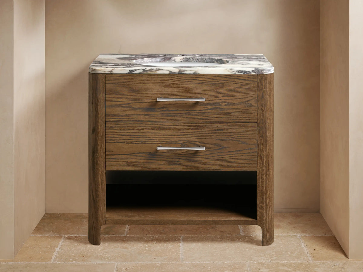Brown oak wood Calista Single Vanity with marble top, silver handles, ceramic sink. Towels placed on lower shelf in a neutral-toned setting.