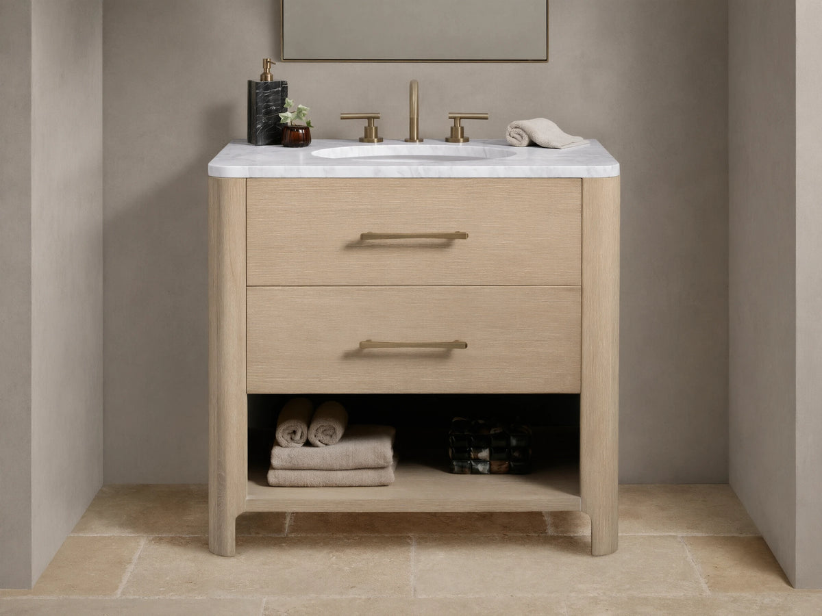 Calista Single Vanity with light oak finish, white marble top, oval sink, dual brass faucets, and storage shelf below.