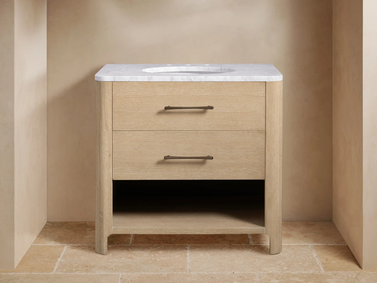 Calista Single Vanity. Light oak wood vanity with white marble top, undermount sink, bronze faucet, two drawers & open shelf with towels.