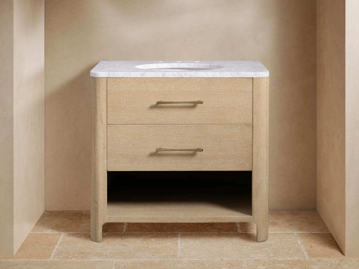 Calista Single Vanity with light oak finish, white marble top, oval sink, dual brass faucets, and storage shelf below.