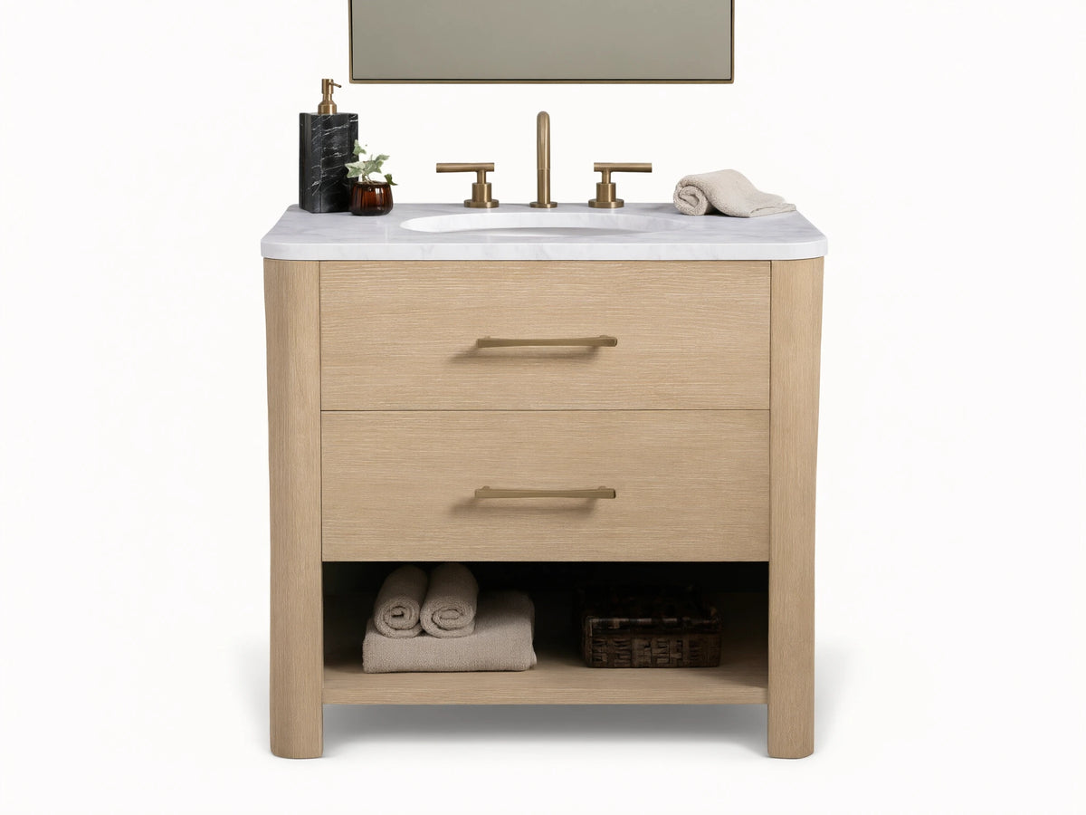 Calista Single Vanity with light oak finish, white marble top, oval sink, dual brass faucets, and storage shelf below.
