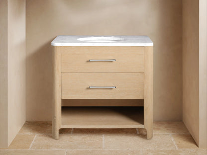 Alt text: Calista Single Vanity with a light oak wood finish and a white marble top. It features two drawers with metal handles, an undermount sink, and an open shelf holding folded towels and jars.