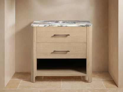 Calista Single Vanity: light oak wood, gray/white marble top, bronze faucet, two drawers with dark pulls. Open shelf with towels.