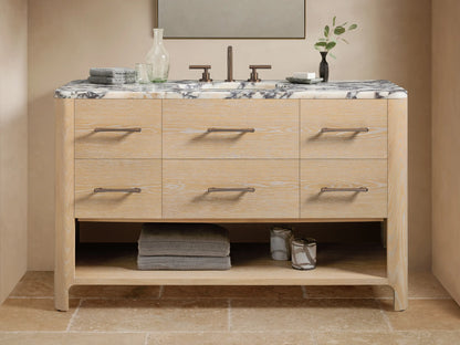 Calista Wide Single Vanity: light oak wood, 6 drawers with dark pulls, white Carrara marble top, dark faucet, open shelf below.