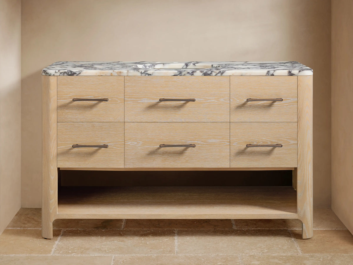 Calista Wide Single Vanity: light oak wood, 6 drawers with dark pulls, white Carrara marble top, dark faucet, open shelf below.