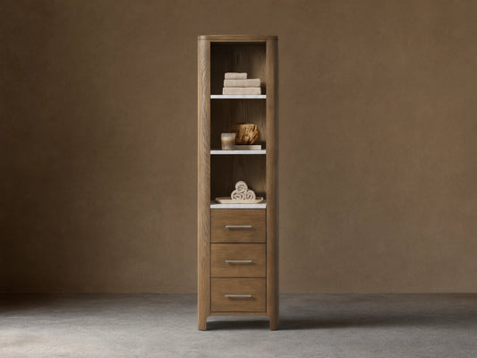 Calista Etagere with oak wood frame, three drawers, and three shelves holding towels. Stone top, against a beige wall.