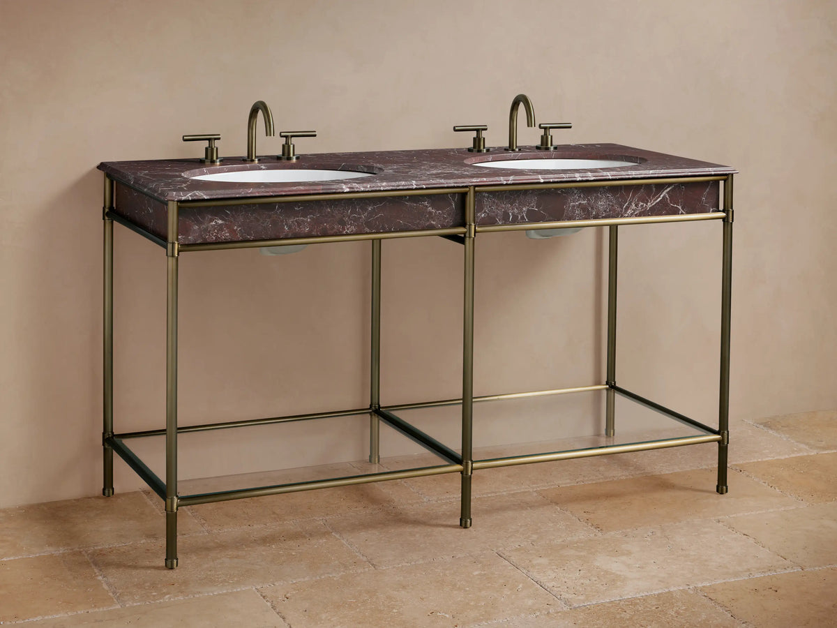 Chelsea Double Washstand. Red Levanto marble top with two white oval sinks. Antique brass frame, lower glass shelf, against a tan wall.