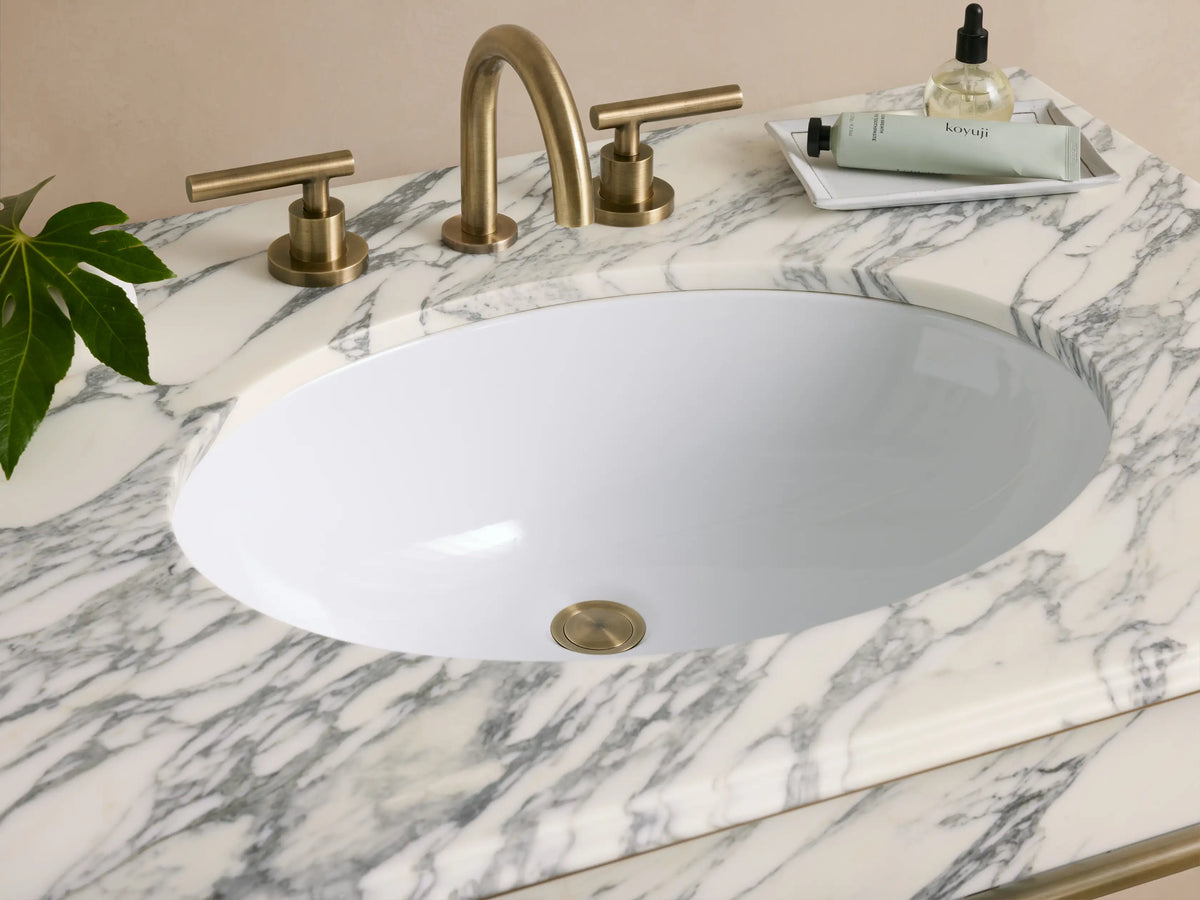 Chelsea Single Washstand with white marble top, oval sink, and antique brass frame. Bottom glass shelf, against a beige wall.