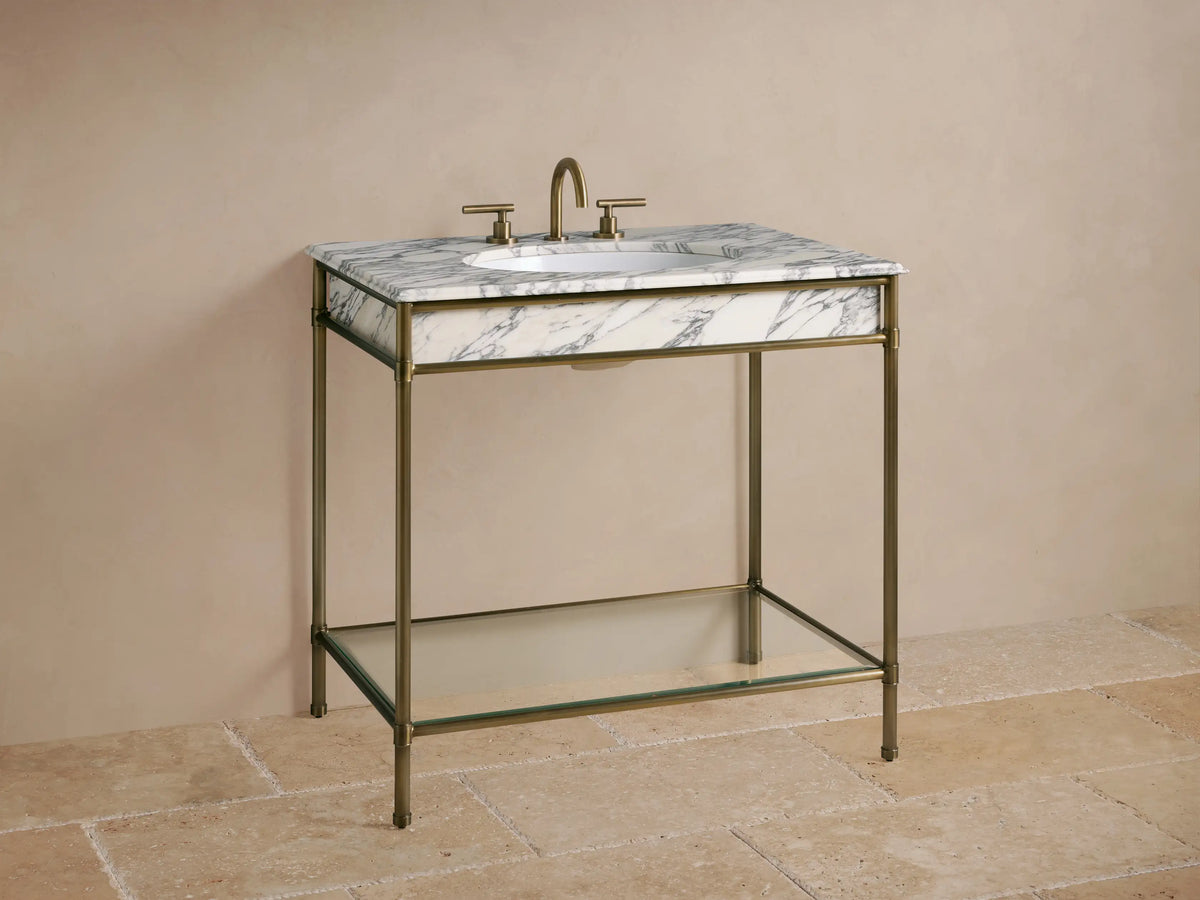 Close-up of Chelsea Single Washstand in Antique Brass, with a glass shelf, sponge, vase, and towels on a stone floor.