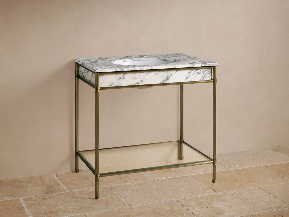 Chelsea Single Washstand with antique brass frame, white marble top with grey veins, oval sink, brass faucet, and lower glass shelf.