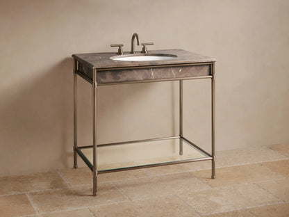 Close-up of Chelsea Single Washstand's lower glass shelf with metallic frame. Marble tray, folded towels, dark vase, grey box, on stone floor.