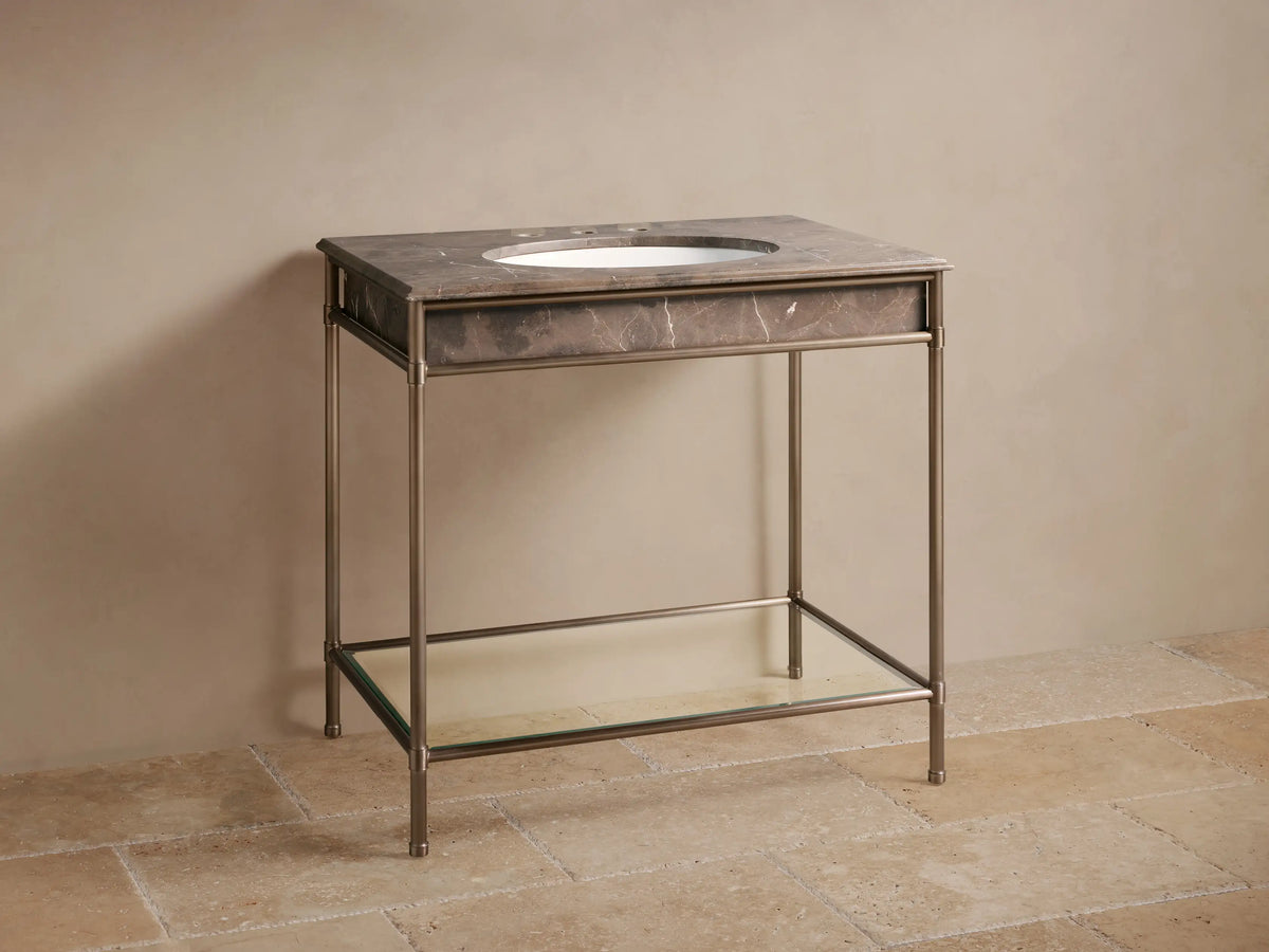 Chelsea Single Washstand. Dark marbled top with white oval sink, bronze faucet, aged metal frame, glass shelf, against beige wall.