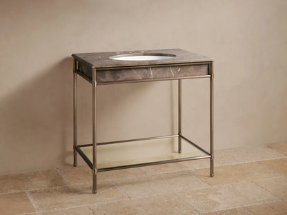 Chelsea Single Washstand. Dark marbled top with white oval sink, bronze faucet, aged metal frame, glass shelf, against beige wall.