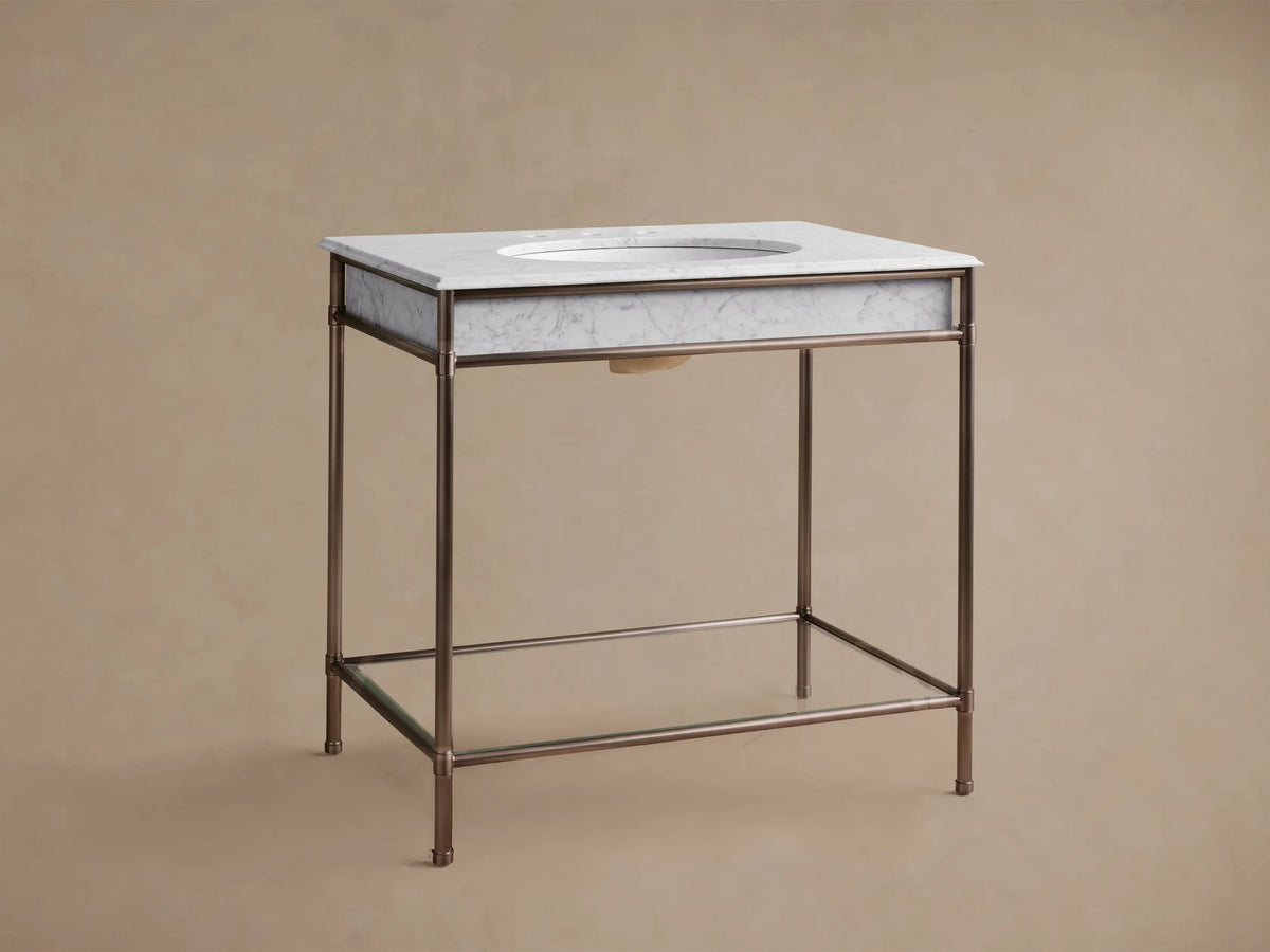 Chelsea Single Washstand, white marble top with oval sink and dark faucet, antique bronze metal frame, and glass shelf.