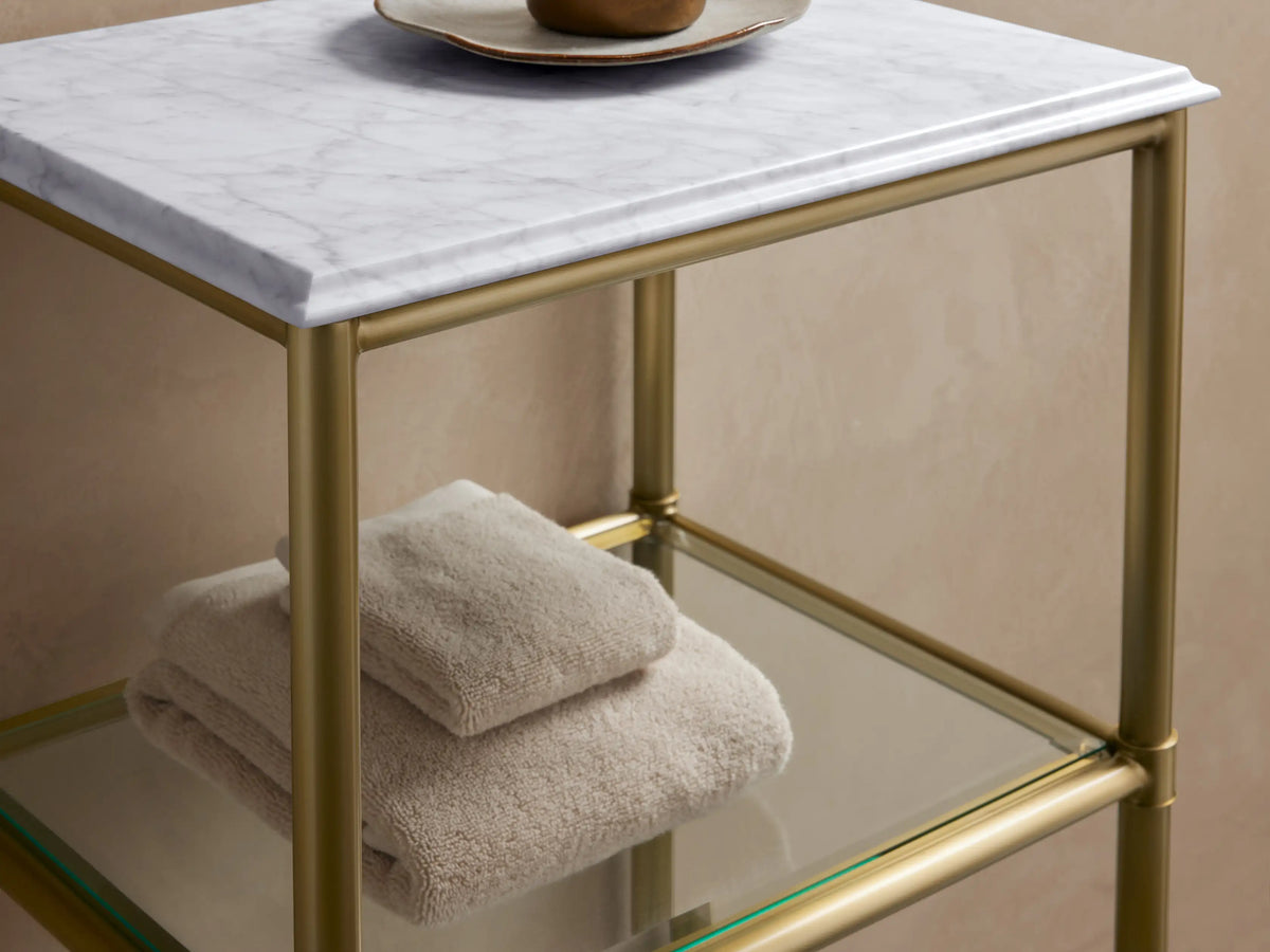 Chelsea Short Etagere with antique brass frame, Carrara marble top, and two glass shelves. Positioned against a neutral wall.