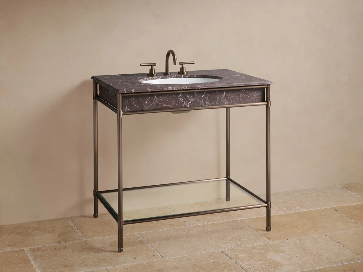 Chelsea Single Washstand's lower shelf: antique bronze frame, glass, light beige towels, dark vase, and box on stone floor.