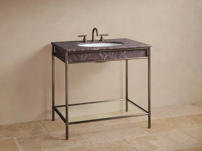 Chelsea Single Washstand's lower shelf: antique bronze frame, glass, light beige towels, dark vase, and box on stone floor.