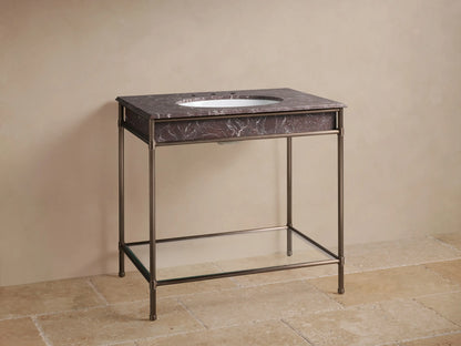 Chelsea Single Washstand with antique bronze frame, dark veined marble top, oval white sink, dark faucet, & clear glass shelf.