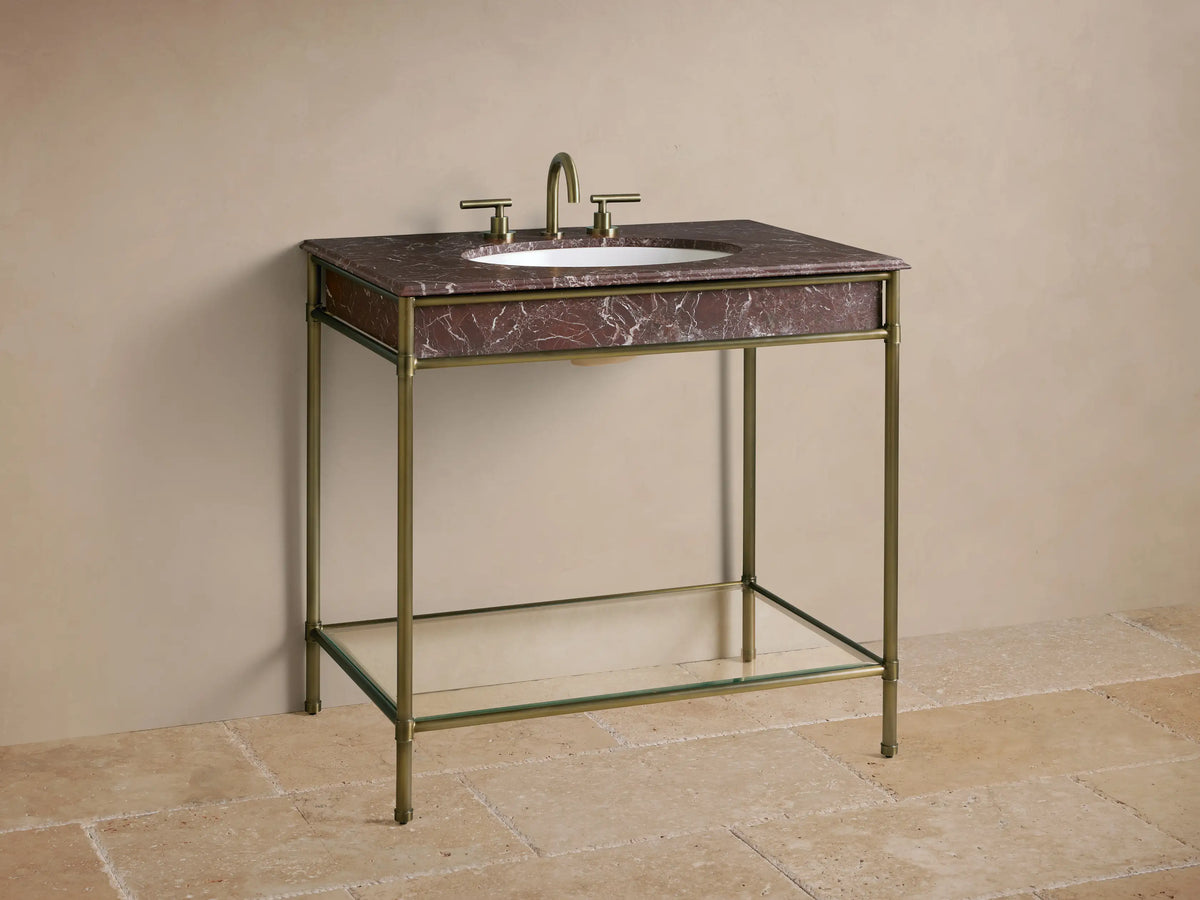 Close-up of Chelsea Single Washstand's antique brass frame and clear glass shelf, with soap, candle, and towels, on stone floor.