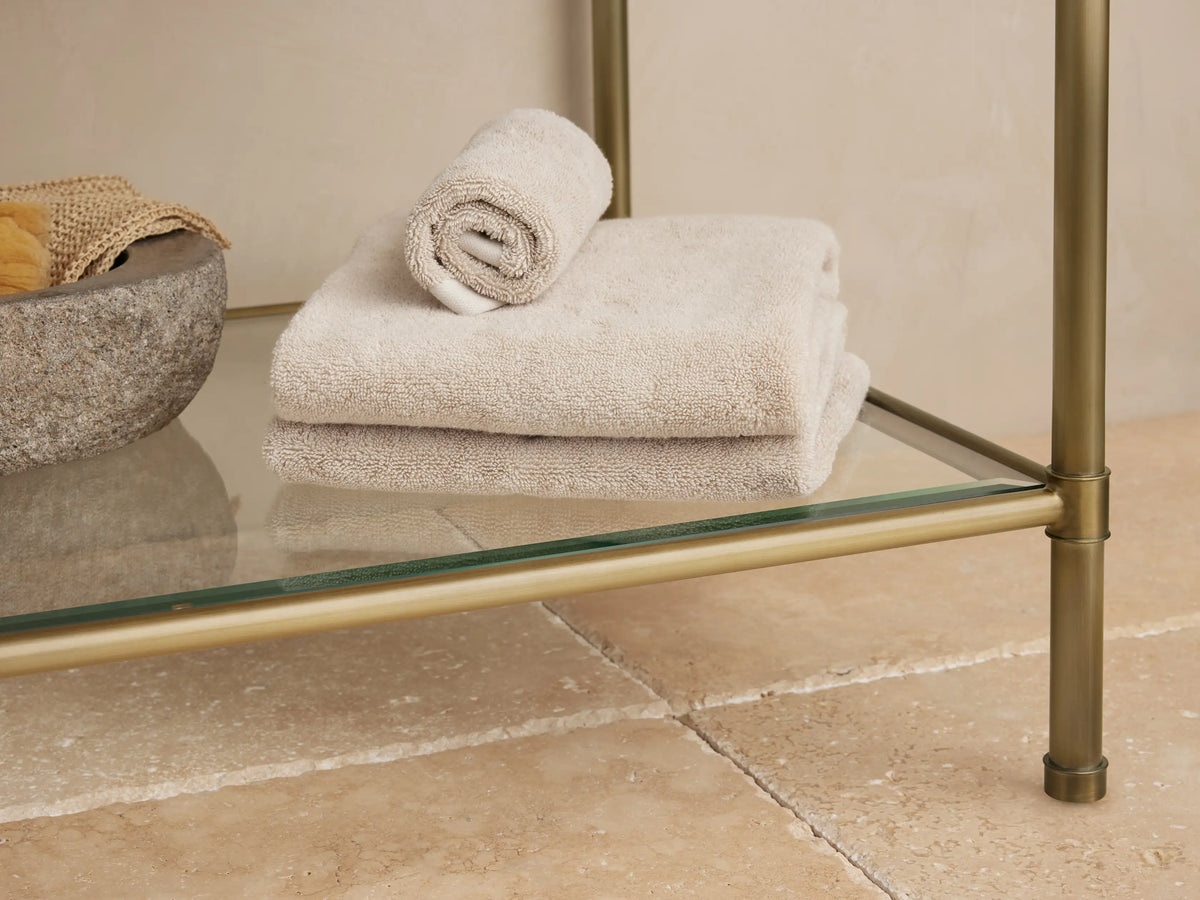 Chelsea Single Washstand's antique brass frame & glass shelf, holding a sponge, driftwood, & towels in a stone bowl.