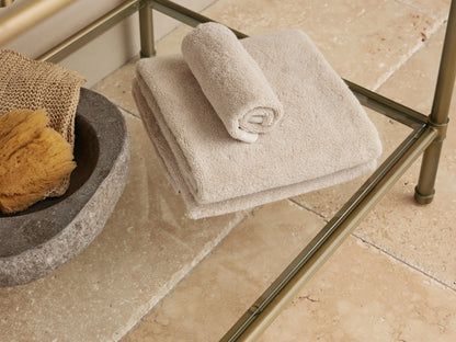Close-up of Chelsea washstand's lower glass shelf with brass frame, holding light beige towels & stone bowl on tan tiled floor.