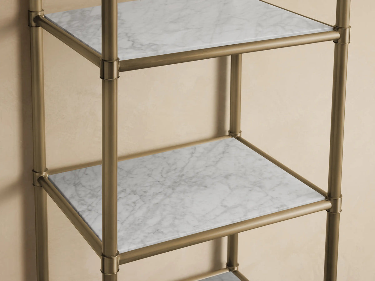 Chelsea Tall Etagere with a stainless steel frame in antique brass finish and five white stone shelves against a beige wall.