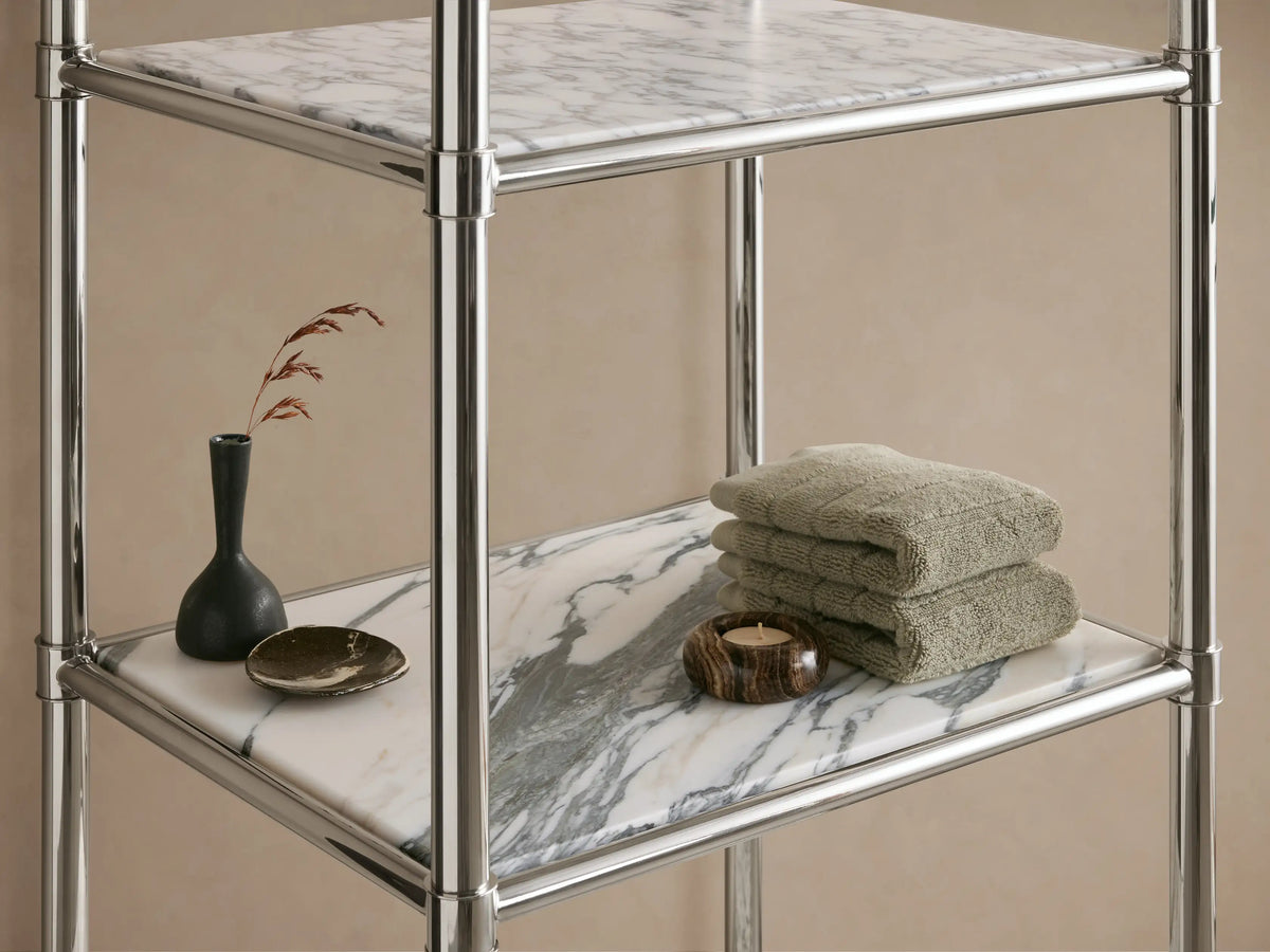 Chelsea Tall Etagere with a shiny polished nickel frame and 5 white Carrara marble shelves, against a beige wall.