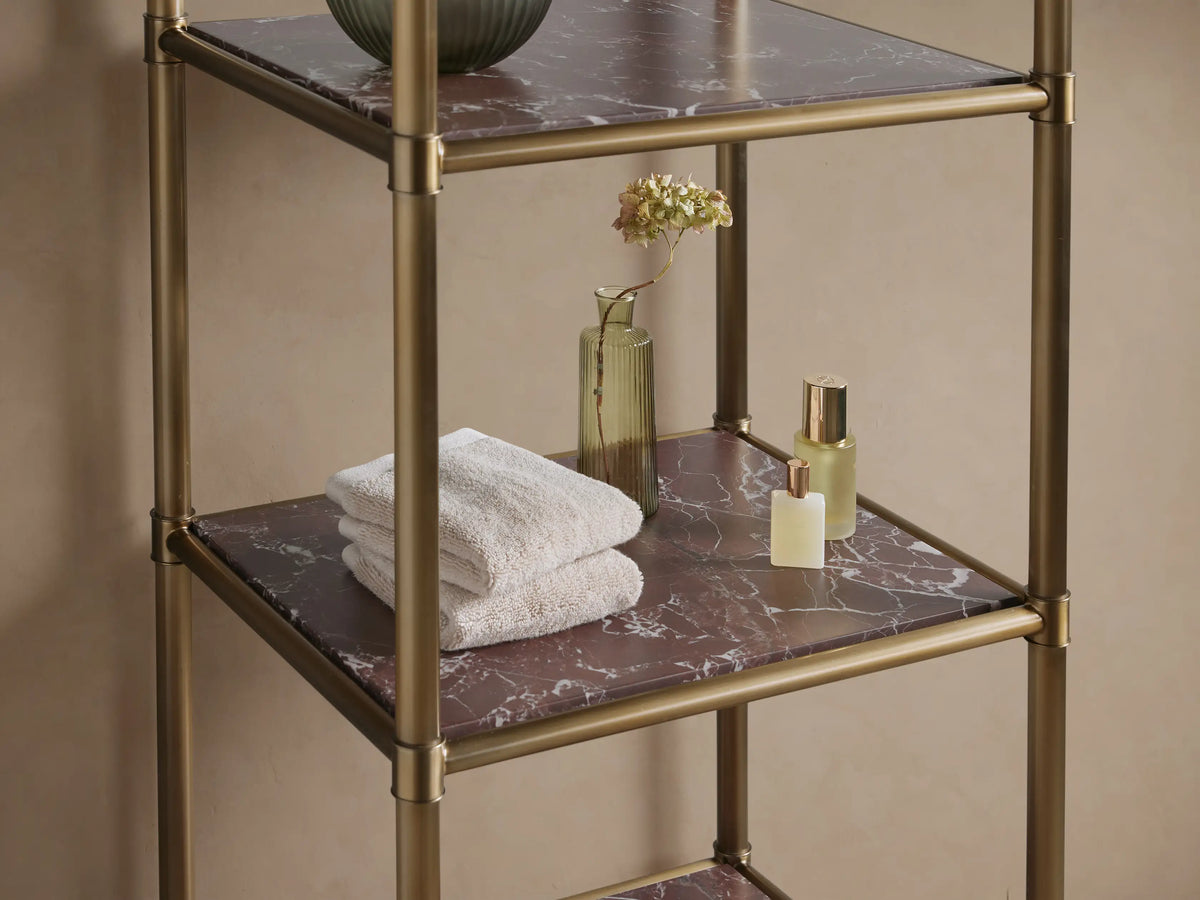 Chelsea Tall Etagere with an antique brass frame and 5 dark, veined marble shelves, standing against a beige wall on stone tiles.