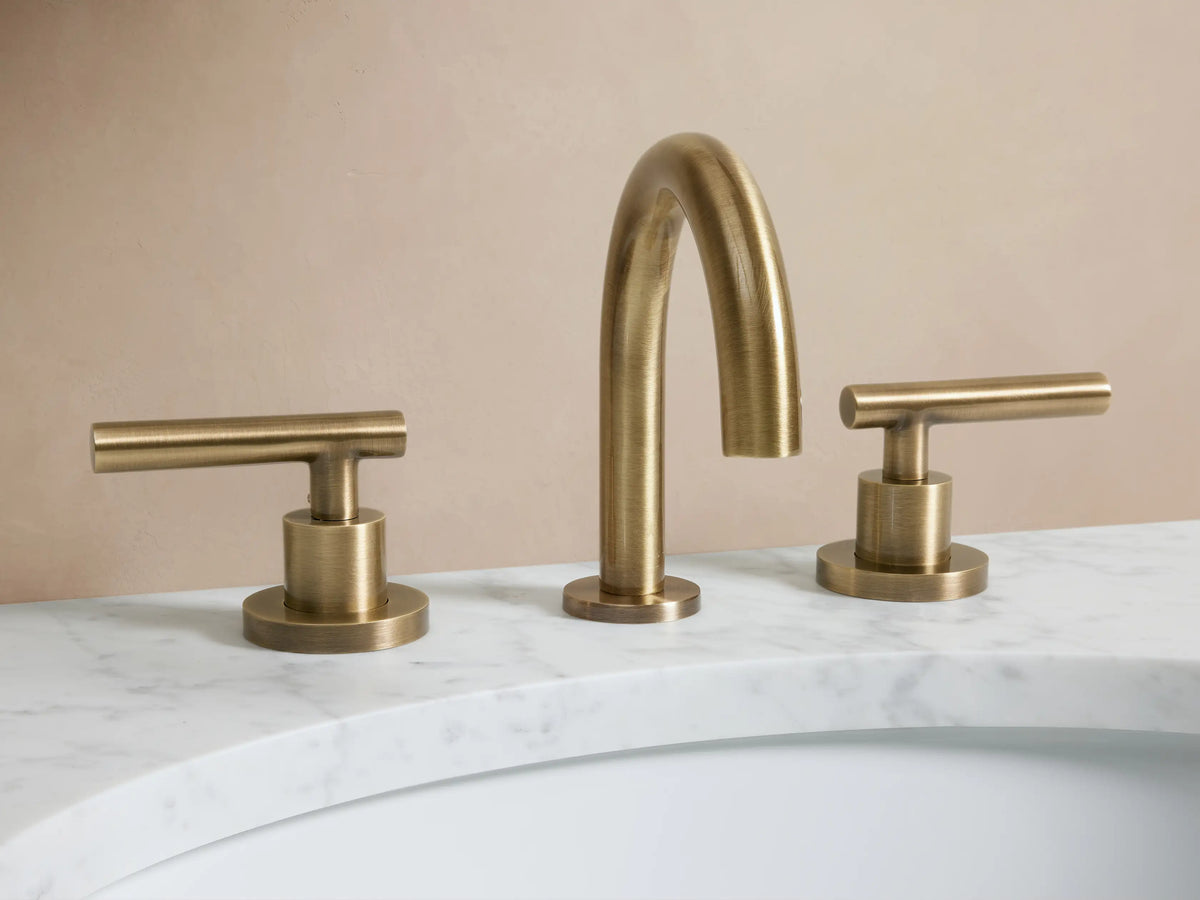 Crewe Smooth Faucet in antique brass, featuring a central curved spout with two cylindrical handles, mounted on a white countertop.