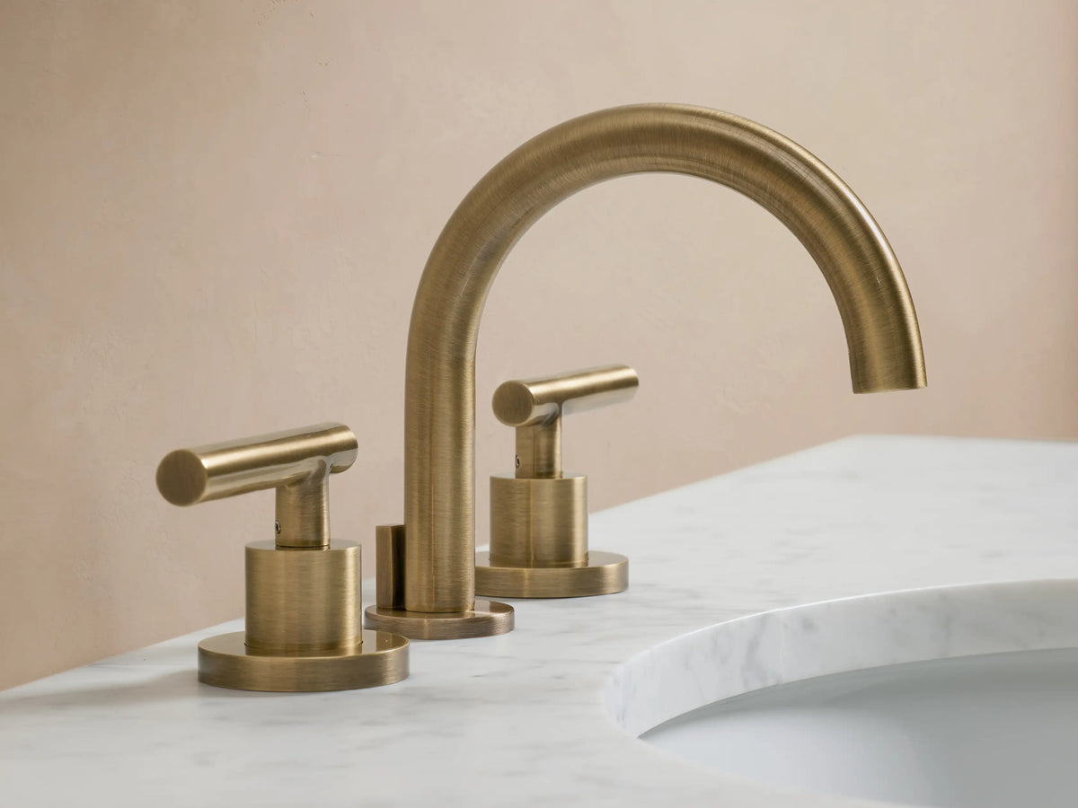 Crewe Smooth Faucet in antique brass, featuring a central curved spout with two cylindrical handles, mounted on a white countertop.
