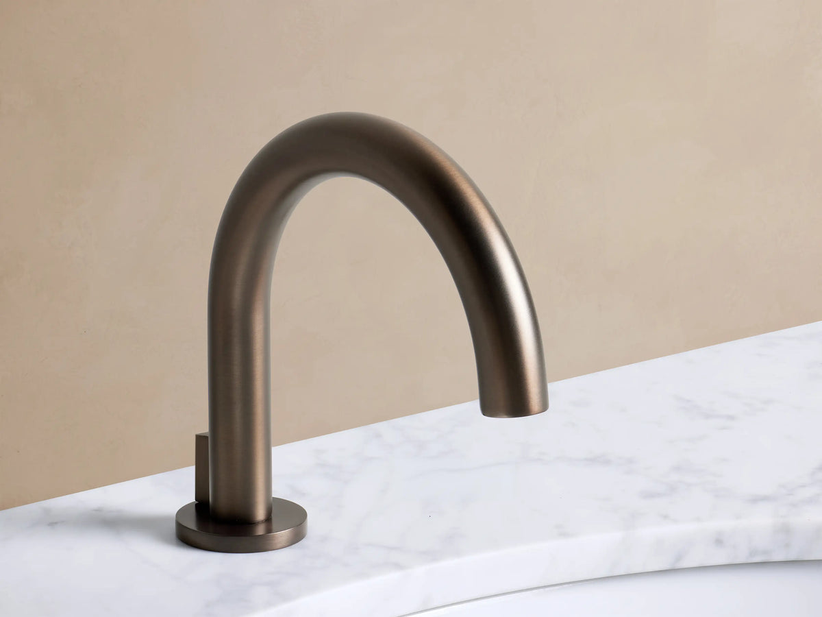 Crewe Smooth Faucet in Bronze. Deck-mounted widespread bronze faucet with curved spout and lever handles on a white marble sink.