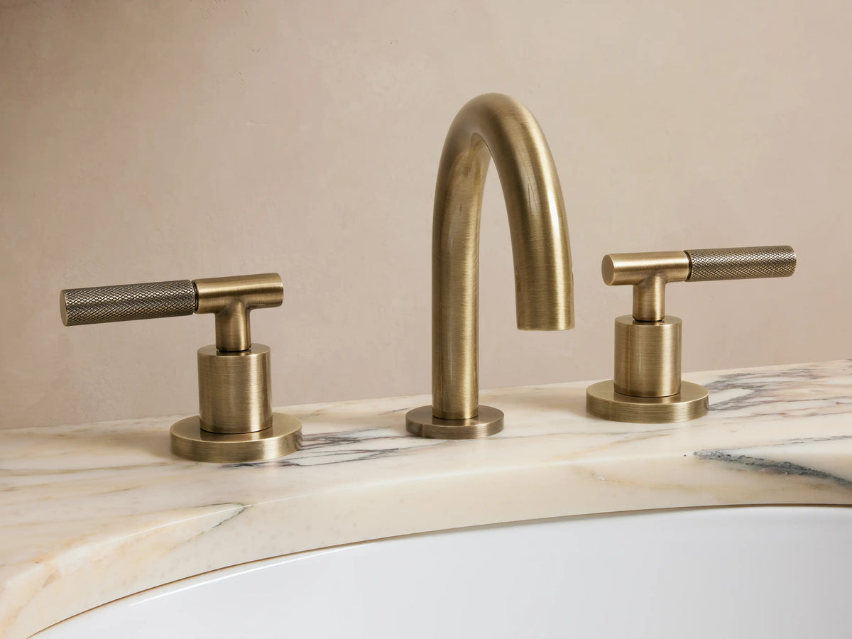 Crewe Knurled Faucet in antique brass finish, featuring a curved spout and two textured handles set on a stone countertop.