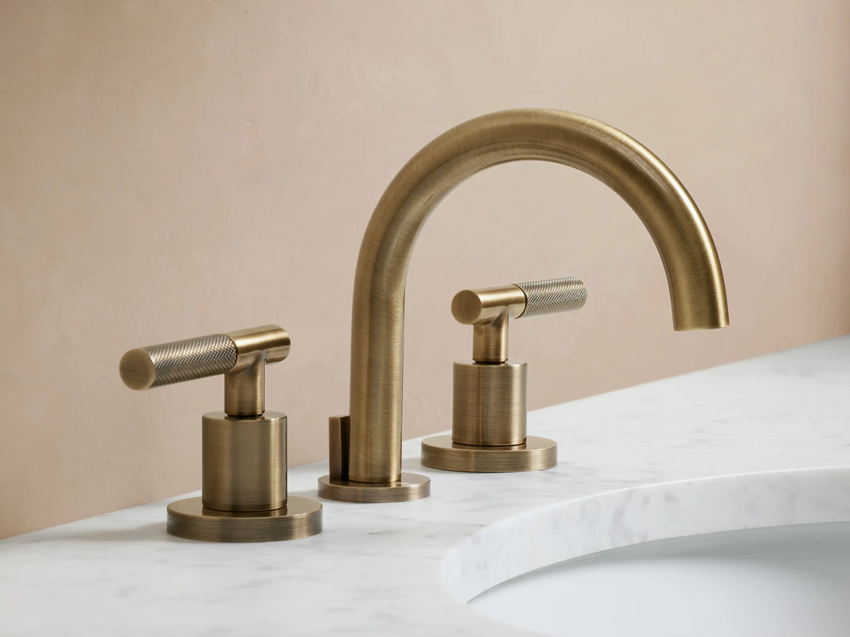 Crewe Knurled Faucet in antique brass finish shown installed on a marble countertop with two side handles and curved spout.